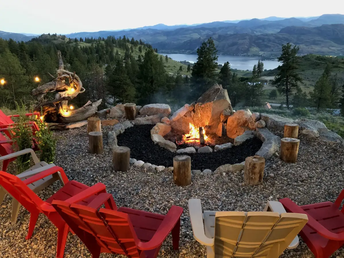 Summit Retreat stone fire pit with colorful chairs, log stools, and driftwood art at dusk
